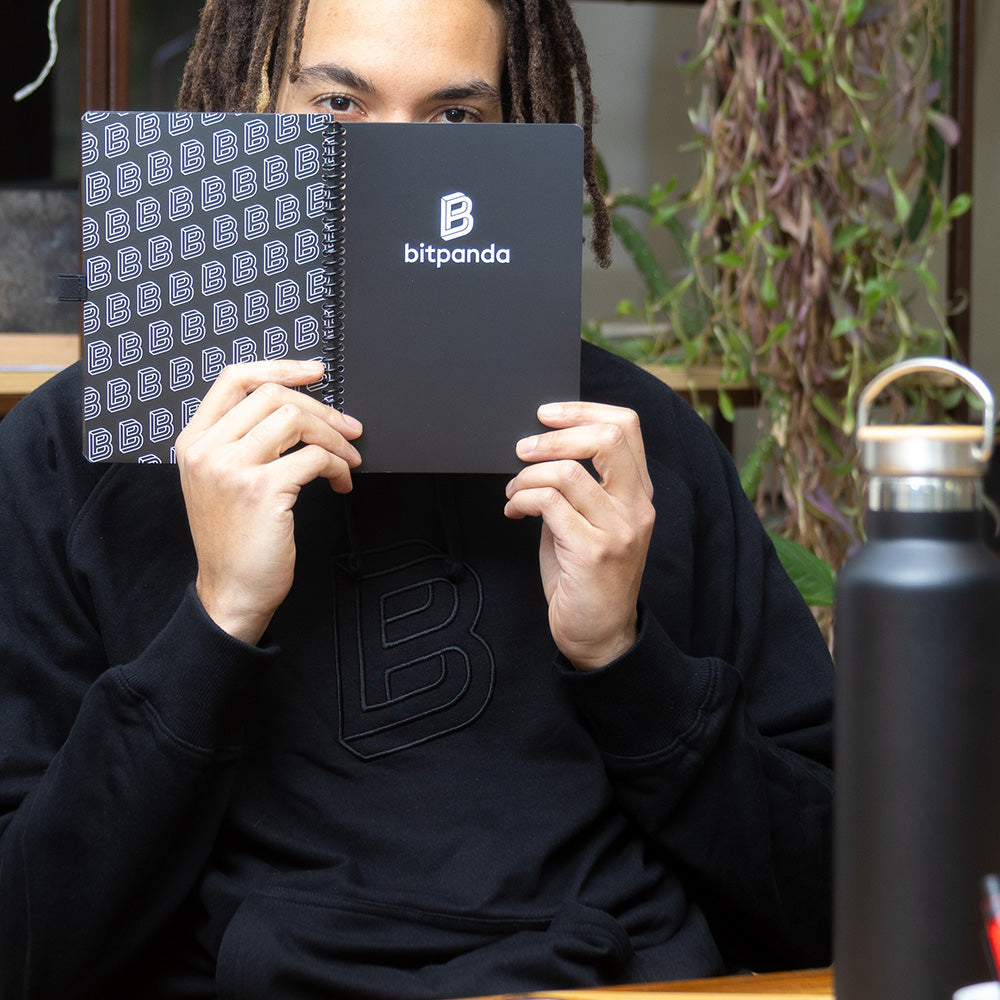 Hände halten einen schwarzen Hoodie mit Bitpanda Logo neben einer Pflanze und einer Trinkflasche, Referenz für individuell bedruckte Firmenkleidung