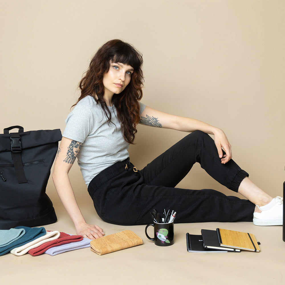 Frau in hellblauem Oberteil und dunkler Hose sitzt lässig auf dem Boden neben einer schwarzen Tasche, Lifestyle Aufnahme für nachhaltige Corporate Fashion