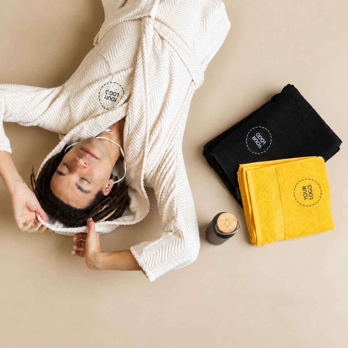 Person mit cremefarbenem Bademantel liegt neben schwarzem und gelbem Handtuch mit Your Logo Platzhalter und Trinkflasche, Mitarbeitergeschenke für Hotellerie und Wellness