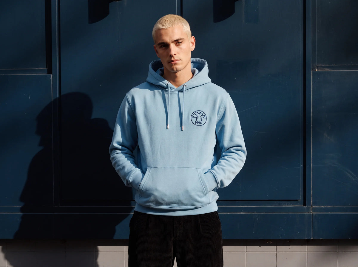 Mann trägt einen hellblauen Hoodie mit gesticktem MMC-Baumlogo auf der Brust – Beispiel für individuell bestickte Mitarbeiter-Hoodies.