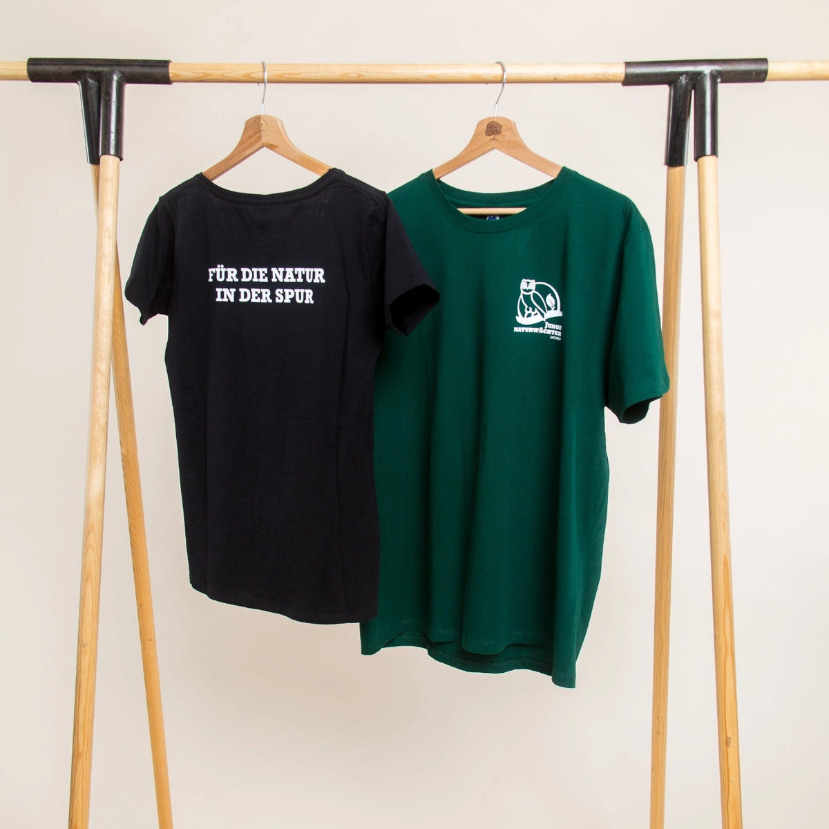 Zwei bedruckte T-Shirts in Schwarz und Grün mit Vereinslogo "Junge Naturwächter Sachsen" und Spruch "Für die Natur in der Spur", Beispiel für individuelles Vereins-Merchandise.
